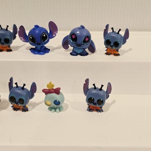 May include: A collection of small, blue Stitch figurines with large eyes and distinctive ears. Some figures have orange accents, while one has a bow and a yellow crown. These collectible toys are from the popular animated movie.