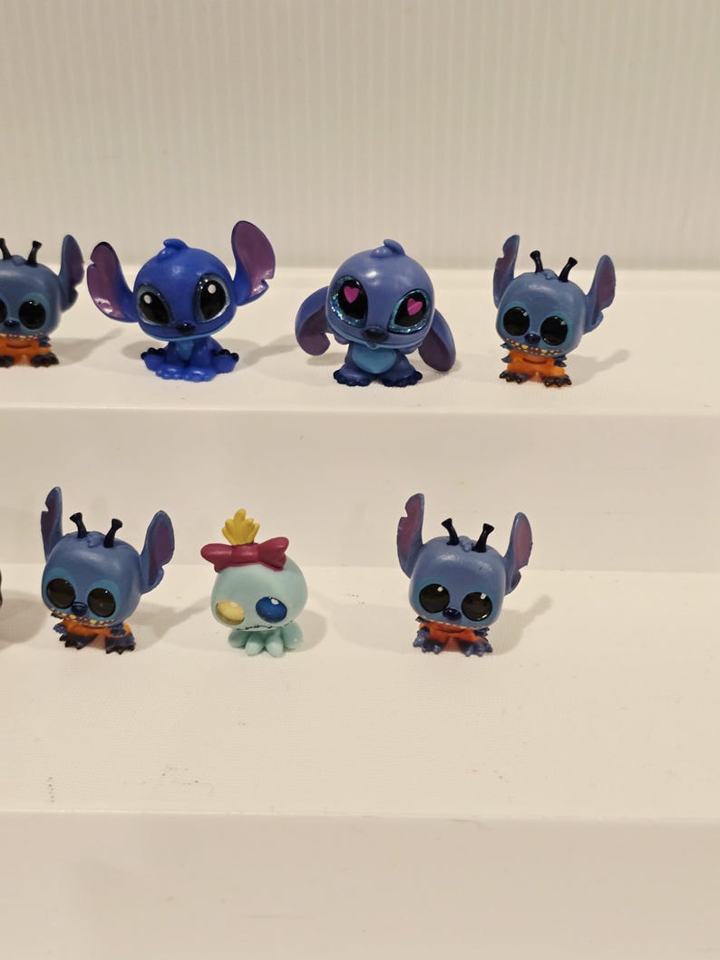 May include: A collection of small, blue Stitch figurines with large eyes and distinctive ears. Some figures have orange accents, while one has a bow and a yellow crown. These collectible toys are from the popular animated movie.