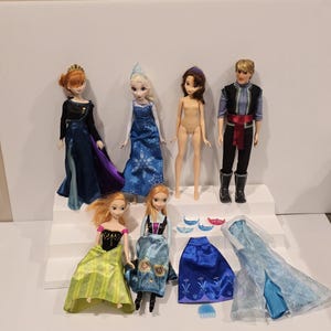May include: A collection of Disney Frozen dolls and accessories. The dolls include Anna, Elsa, Kristoff, and other characters. Accessories include dresses, crowns, and a comb. The dolls are posed on a white stepped platform.