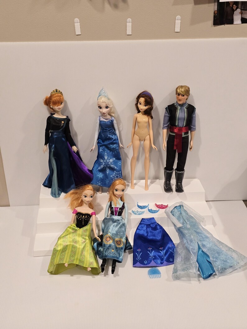 May include: A collection of Disney Frozen dolls and accessories. The dolls include Anna, Elsa, Kristoff, and other characters. Accessories include dresses, crowns, and a comb. The dolls are posed on a white stepped platform.