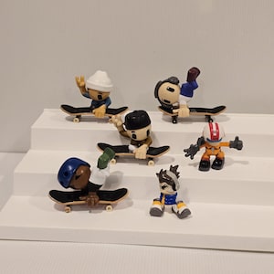 May include: A collection of miniature skateboarder figurines in various poses. Each figure is on a small skateboard with black decks and yellow wheels. The skaters wear different outfits, including helmets and jackets, and are in mid-trick positions.