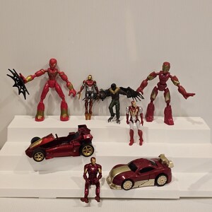 May include: A collection of red and gold action figures and toy cars. The figures include Spider-Man, Iron Man, and Vulture. The toy cars are red and gold. The toys are arranged on white steps.