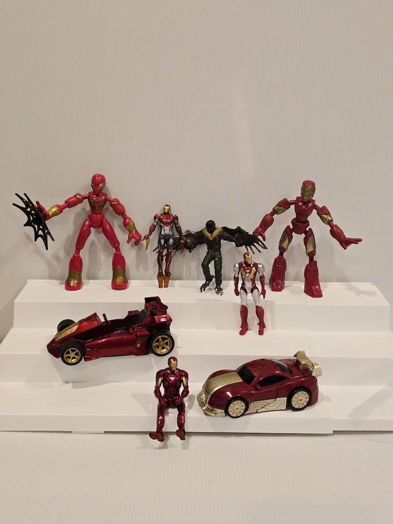 May include: A collection of red and gold action figures and toy cars. The figures include Spider-Man, Iron Man, and Vulture. The toy cars are red and gold. The toys are arranged on white steps.