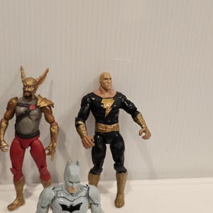 May include: Three action figures of DC Comics characters. The figures include Hawkman in red and gold, Black Adam in black and gold, and Batman in gray and black. The figures are standing on a white surface.