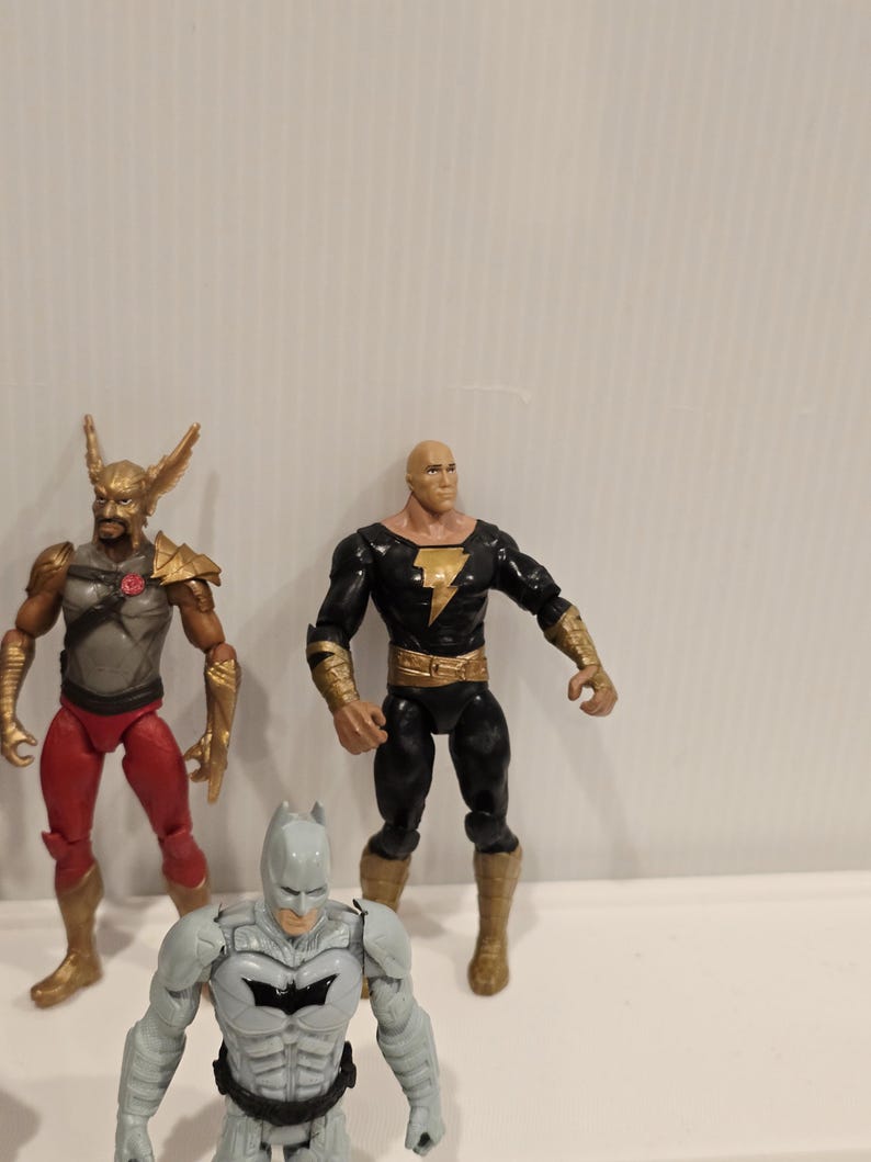 May include: Three action figures of DC Comics characters. The figures include Hawkman in red and gold, Black Adam in black and gold, and Batman in gray and black. The figures are standing on a white surface.