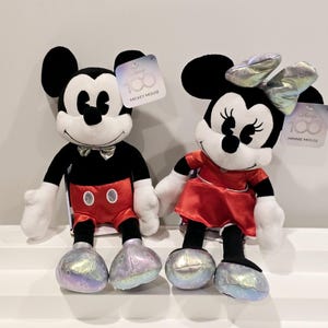 以下が含まれることがあります： 黒と白のミッキーマウスのぬいぐるみと、赤と白のミニーマウスのぬいぐるみ。両方のぬいぐるみは、それぞれ赤いショートパンツと赤いドレスを着ています。両方のぬいぐるみは、キラキラと虹色の仕上げが施されており、ミッキーマウスの100周年を祝っています。