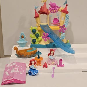K&ouml;nnte beinhalten: Ariel's Sea Castle Spielset mit einem gelb-roten Schloss, einer blauen Rutsche und verschiedenen Figuren. Das Set enth&auml;lt ein Boot und Figuren von Arielle in blauen und rosa Kleidern. Ein rosa Anleitungsheft ist ebenfalls enthalten.