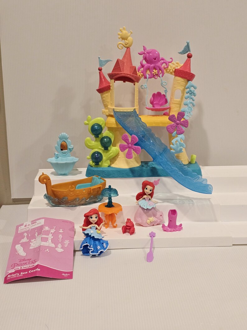 K&ouml;nnte beinhalten: Ariel's Sea Castle Spielset mit einem gelb-roten Schloss, einer blauen Rutsche und verschiedenen Figuren. Das Set enth&auml;lt ein Boot und Figuren von Arielle in blauen und rosa Kleidern. Ein rosa Anleitungsheft ist ebenfalls enthalten.