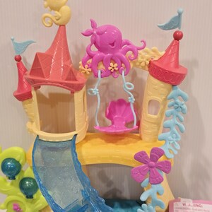 May include: A colorful toy playset featuring a castle design with a slide and swing. The castle has pink and yellow towers, a blue slide, and is decorated with seahorses, an octopus, and flowers. The playset is designed for children.