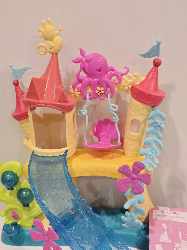 May include: A colorful toy playset featuring a castle design with a slide and swing. The castle has pink and yellow towers, a blue slide, and is decorated with seahorses, an octopus, and flowers. The playset is designed for children.