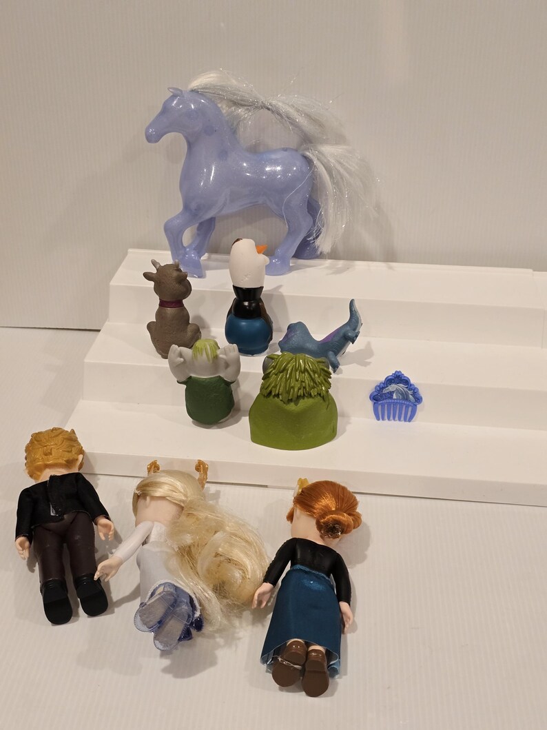 May include: A collection of toy figures from the animated movie "Frozen." The set includes a light purple horse, a reindeer, a snowman, and three dolls. The dolls have blonde and red hair, and are wearing various outfits.