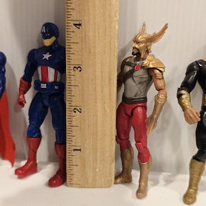 May include: Action figures of superheroes, including Captain America, and Hawkman. The Captain America figure is blue, red, and white, and the Hawkman figure is red, gold, and gray. A wooden ruler is next to the figures, measuring approximately 4 inches.