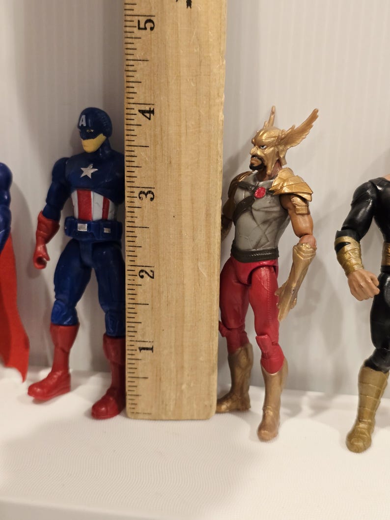 May include: Action figures of superheroes, including Captain America, and Hawkman. The Captain America figure is blue, red, and white, and the Hawkman figure is red, gold, and gray. A wooden ruler is next to the figures, measuring approximately 4 inches.