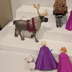 May include: A collection of Frozen-themed figurines. The set includes a reindeer with large antlers, a snowman, and figures of Elsa and Anna in purple and pink capes. Other figures are also visible.