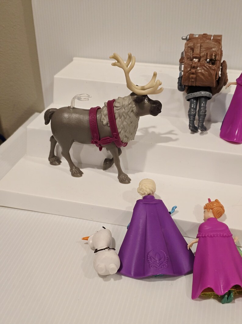 May include: A collection of Frozen-themed figurines. The set includes a reindeer with large antlers, a snowman, and figures of Elsa and Anna in purple and pink capes. Other figures are also visible.