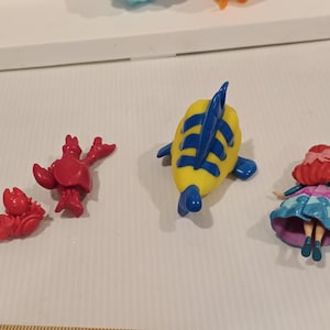 May include: A collection of small plastic figurines featuring characters from a popular animated film. The figures include a red crab, a yellow and blue fish, and a doll with red hair and a blue dress.