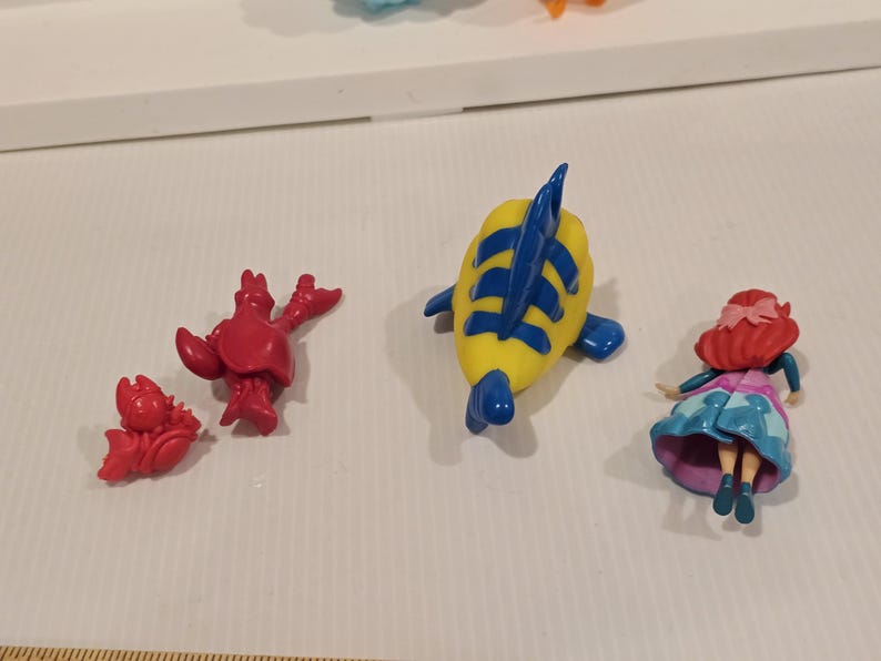 May include: A collection of small plastic figurines featuring characters from a popular animated film. The figures include a red crab, a yellow and blue fish, and a doll with red hair and a blue dress.
