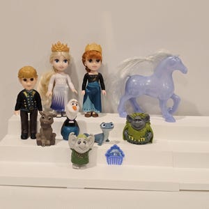 May include: A collection of Frozen-themed figurines, including dolls of Anna and Elsa wearing crowns, Kristoff, Olaf, Sven, a horse, and other characters. The figures are arranged on a white stepped display.