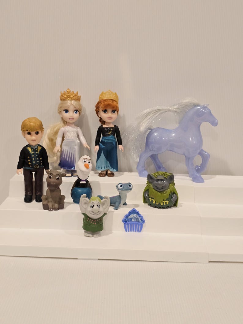 May include: A collection of Frozen-themed figurines, including dolls of Anna and Elsa wearing crowns, Kristoff, Olaf, Sven, a horse, and other characters. The figures are arranged on a white stepped display.