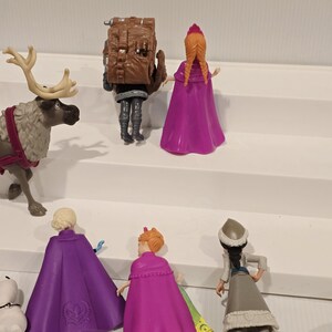 May include: A collection of Frozen character figurines, including Elsa, Anna, Kristoff, and Sven the reindeer. The figures are in various poses, with vibrant colors and detailed designs. The set includes a small snowman figurine.