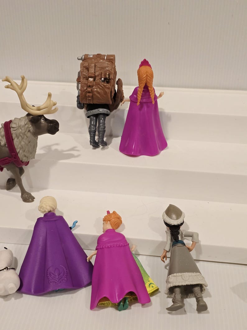 May include: A collection of Frozen character figurines, including Elsa, Anna, Kristoff, and Sven the reindeer. The figures are in various poses, with vibrant colors and detailed designs. The set includes a small snowman figurine.