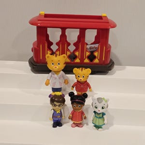 May include: A set of Daniel Tiger's Neighborhood figurines, including Daniel Tiger, Katerina Kittycat, O the Owl, and others. The figurines are standing in front of a red trolley car toy.