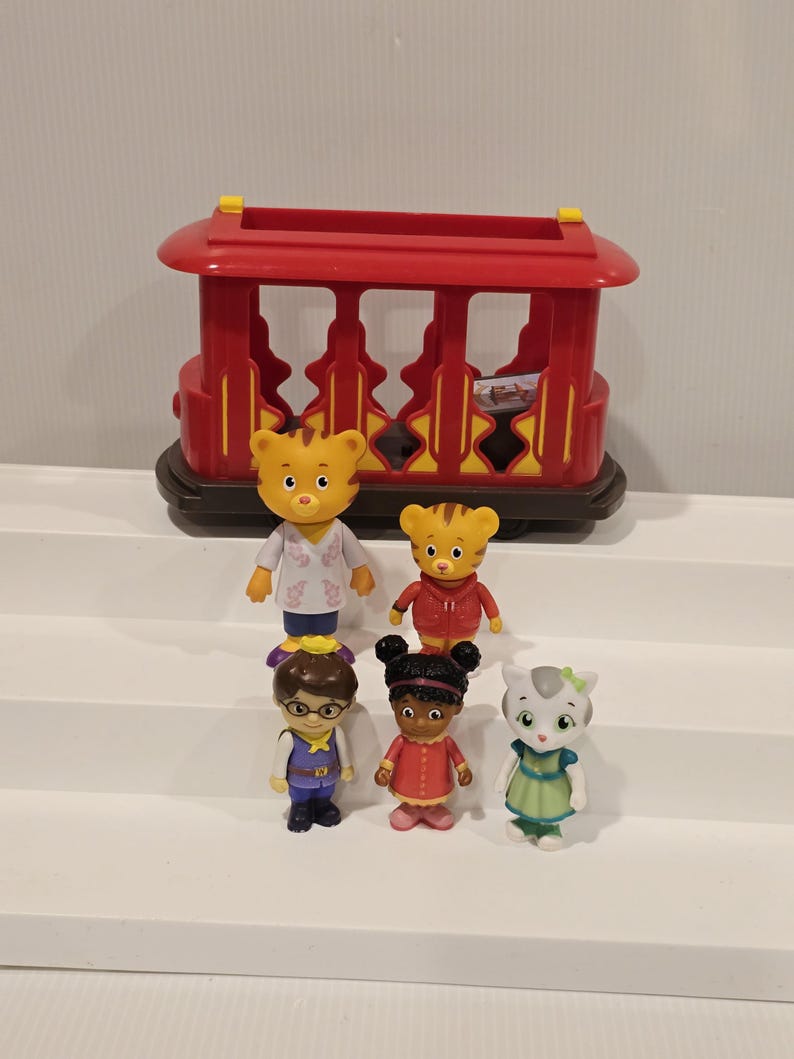 May include: A set of Daniel Tiger's Neighborhood figurines, including Daniel Tiger, Katerina Kittycat, O the Owl, and others. The figurines are standing in front of a red trolley car toy.