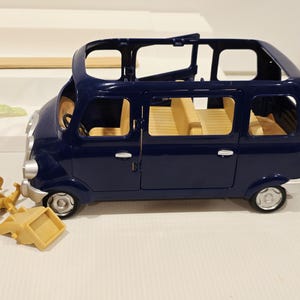 May include: A dark blue toy van with tan interior seating and silver-colored wheels. The van has an open roof and side doors. Several tan-colored chairs are scattered near the van. A light blue ring and a green object are in the background.