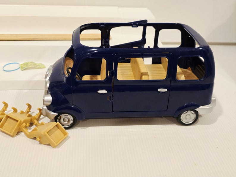 May include: A dark blue toy van with tan interior seating and silver-colored wheels. The van has an open roof and side doors. Several tan-colored chairs are scattered near the van. A light blue ring and a green object are in the background.