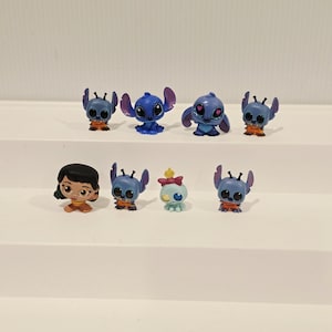 May include: A collection of small, colorful figurines featuring characters from the animated film Lilo & Stitch. The figurines include Stitch in various poses, along with Lilo and another character. The figures are primarily blue, orange, and brown.