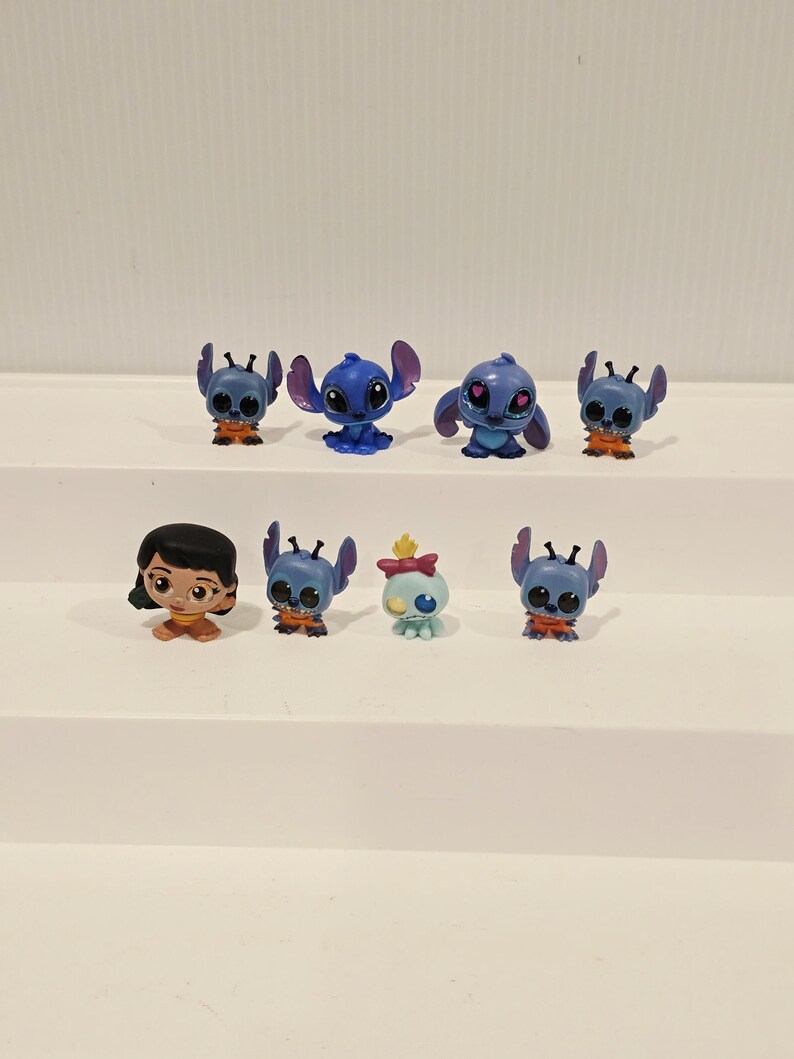 May include: A collection of small, colorful figurines featuring characters from the animated film Lilo & Stitch. The figurines include Stitch in various poses, along with Lilo and another character. The figures are primarily blue, orange, and brown.