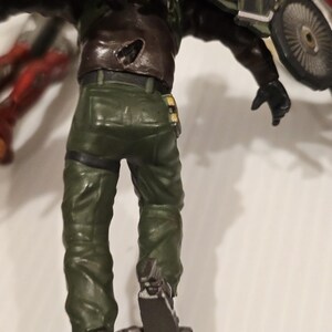 May include: A detailed action figure of a character in a green military-style uniform and brown jacket. The figure has a mechanical device attached to its back and feet. The figure is posed with its back facing the viewer.