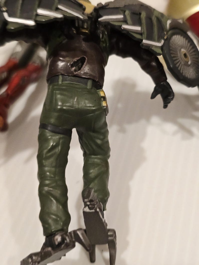 May include: A detailed action figure of a character in a green military-style uniform and brown jacket. The figure has a mechanical device attached to its back and feet. The figure is posed with its back facing the viewer.