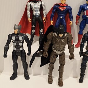 May include: A collection of superhero action figures, including Thor, Superman, Batman, and Captain America. The figures are brightly colored and feature detailed costumes and accessories. The figures are standing on a white surface.