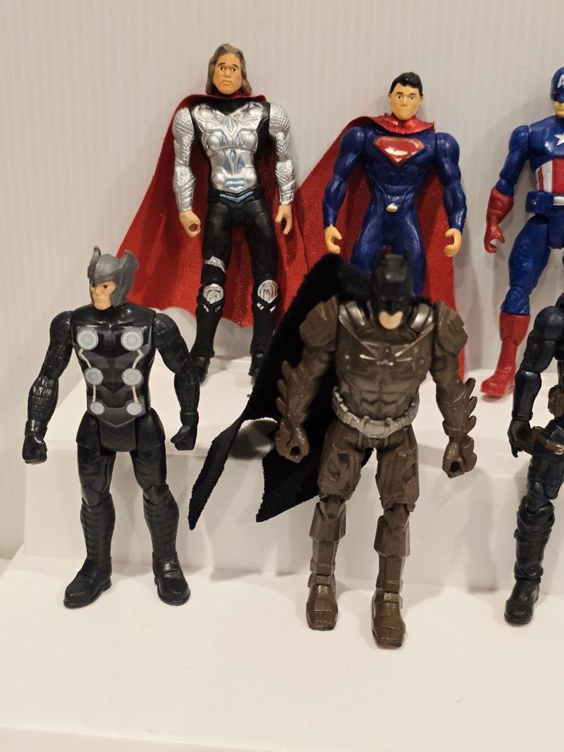 May include: A collection of superhero action figures, including Thor, Superman, Batman, and Captain America. The figures are brightly colored and feature detailed costumes and accessories. The figures are standing on a white surface.