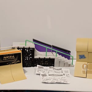 May include: A Tech Deck Mega Halfpipe skate park set. The set includes a halfpipe, stairs, rails, and boxes. The halfpipe is tan and black, with the words "Mega Halfpipe" printed on it. The set also includes instructions and boxes.