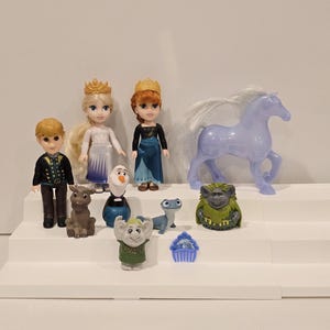May include: A collection of miniature Frozen-themed figurines. The set includes dolls of Elsa, Anna, Kristoff, and Olaf, along with Sven the reindeer, a horse, and other characters. The figures are arranged on a white stepped display.