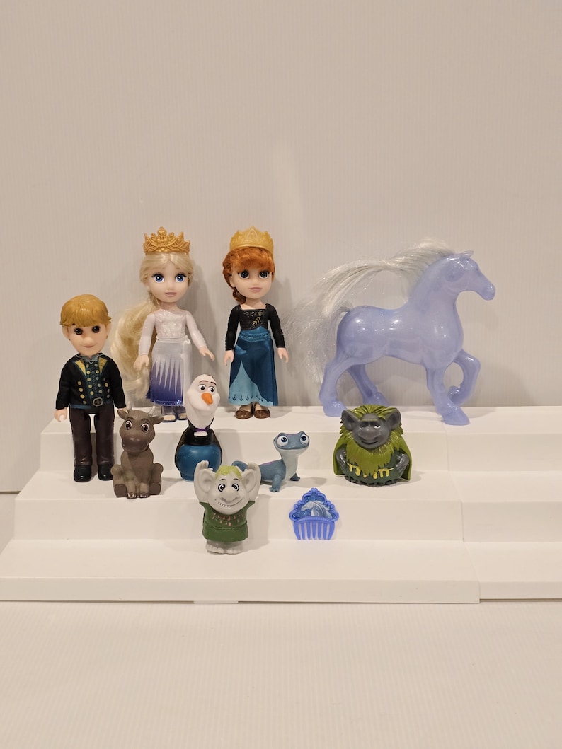 May include: A collection of miniature Frozen-themed figurines. The set includes dolls of Elsa, Anna, Kristoff, and Olaf, along with Sven the reindeer, a horse, and other characters. The figures are arranged on a white stepped display.
