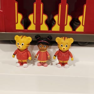 K&ouml;nnte beinhalten: Drei Figuren von Daniel Tiger und einer Freundin stehen vor einem roten Spielzeugzug. Die Daniel Tiger-Figuren sind gelb mit orangefarbenen Streifen und tragen rote Jacken. Die andere Figur ist ein M&auml;dchen in einem roten Kleid.