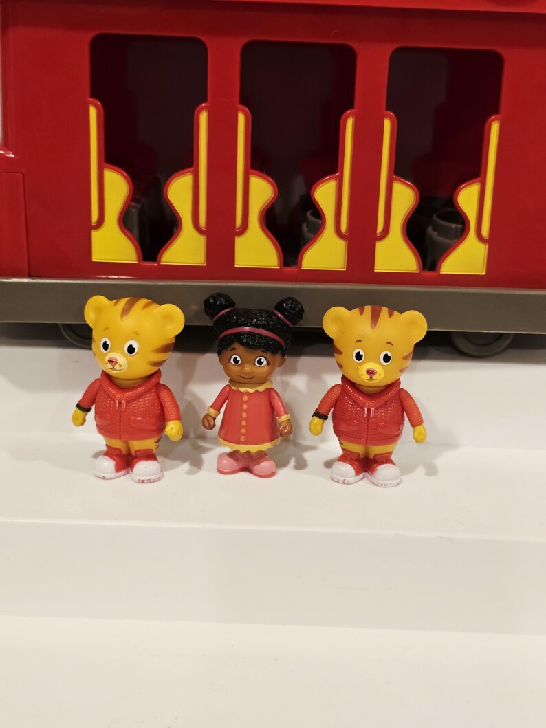 K&ouml;nnte beinhalten: Drei Figuren von Daniel Tiger und einer Freundin stehen vor einem roten Spielzeugzug. Die Daniel Tiger-Figuren sind gelb mit orangefarbenen Streifen und tragen rote Jacken. Die andere Figur ist ein M&auml;dchen in einem roten Kleid.