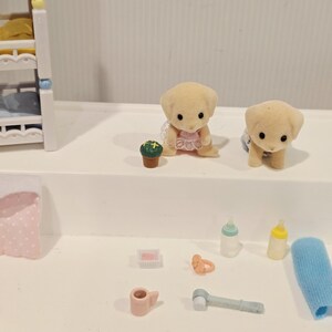 Puede incluir: Colecci&oacute;n de accesorios de juguete en miniatura, que incluye dos figuras de cachorros de color canela, una peque&ntilde;a planta en maceta, biberones, un chupete, un cepillo de dientes y un pa&ntilde;o. En el fondo se ve una cuna en miniatura. Los juguetes est&aacute;n colocados sobre una superficie blanca.