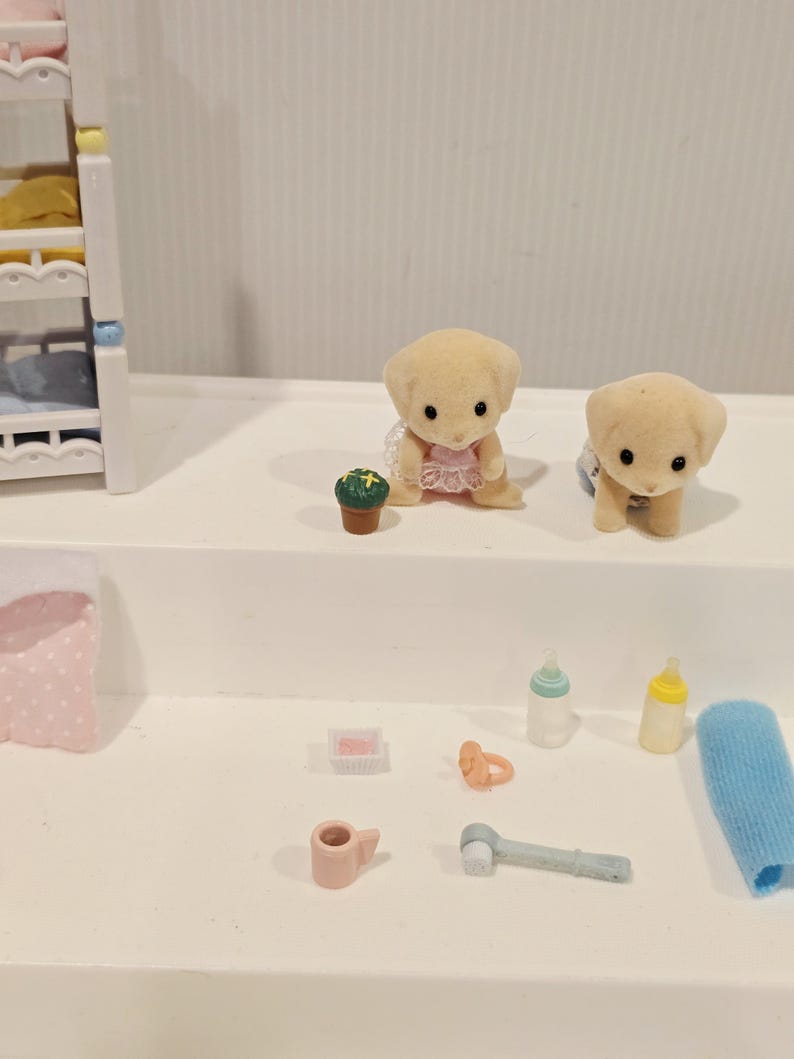 Puede incluir: Colecci&oacute;n de accesorios de juguete en miniatura, que incluye dos figuras de cachorros de color canela, una peque&ntilde;a planta en maceta, biberones, un chupete, un cepillo de dientes y un pa&ntilde;o. En el fondo se ve una cuna en miniatura. Los juguetes est&aacute;n colocados sobre una superficie blanca.