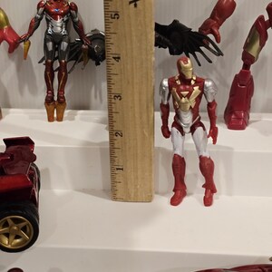 May include: Several Iron Man action figures are displayed, showcasing the iconic superhero in various poses. The figures are primarily red, gold, silver, and black, with detailed designs. A wooden ruler is placed next to the figures.