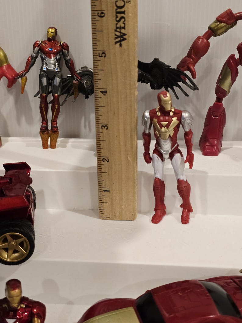 May include: Several Iron Man action figures are displayed, showcasing the iconic superhero in various poses. The figures are primarily red, gold, silver, and black, with detailed designs. A wooden ruler is placed next to the figures.