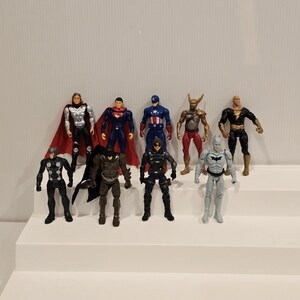 May include: A collection of ten superhero action figures, each with unique costumes and poses. The figures are displayed on a white tiered platform against a plain white background. The figures include Superman, Batman, and Captain America.