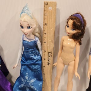 May include: Two dolls, one in a blue gown with snowflake patterns and a silver tiara, and a nude doll with brown hair and a purple headband. A wooden ruler is positioned between them.
