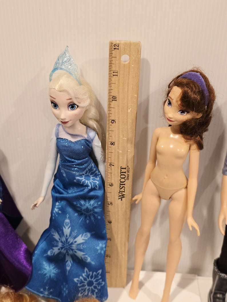 May include: Two dolls, one in a blue gown with snowflake patterns and a silver tiara, and a nude doll with brown hair and a purple headband. A wooden ruler is positioned between them.