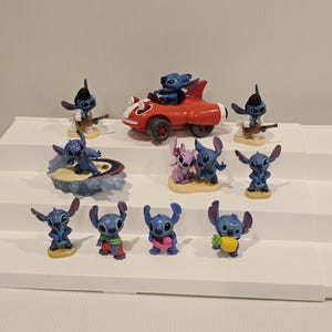 May include: A collection of blue Stitch figurines in various poses. One figurine is driving a red car, others are playing a guitar, surfing, or holding a pineapple. The figurines are on a white shelf.