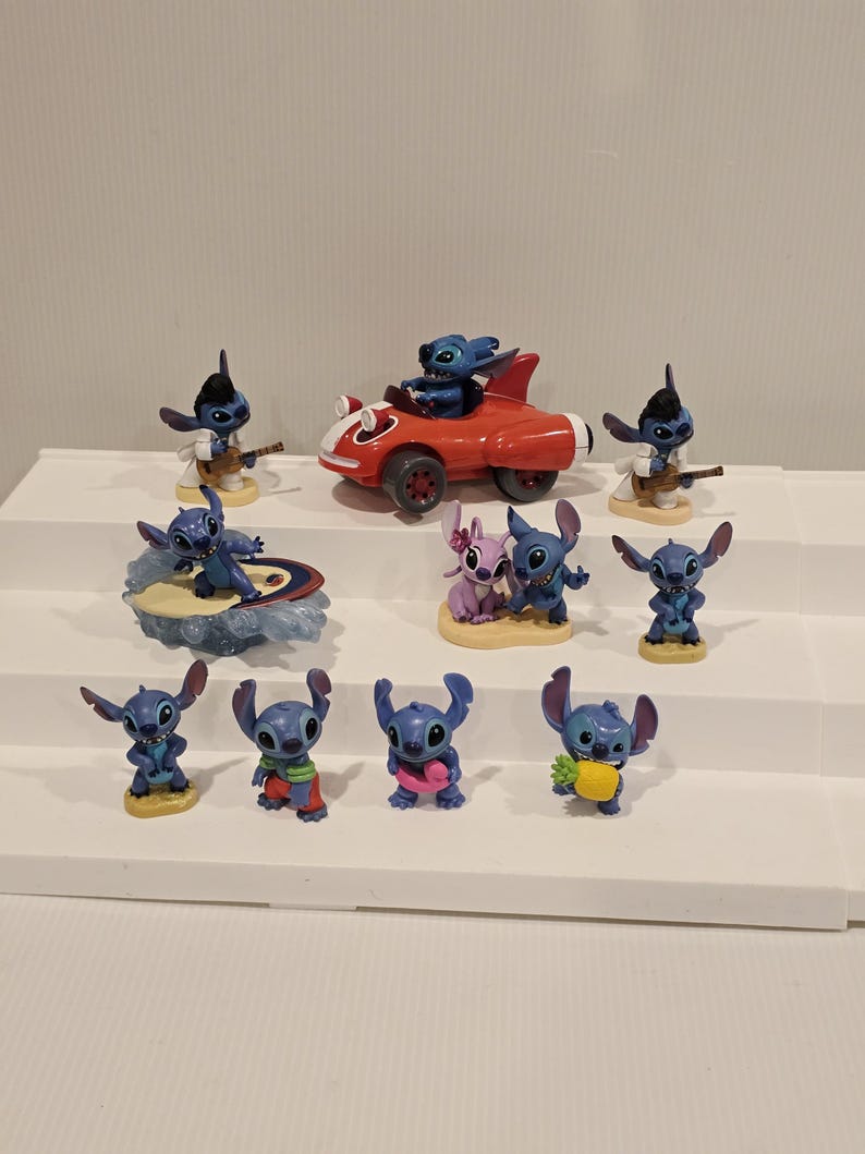 May include: A collection of blue Stitch figurines in various poses. One figurine is driving a red car, others are playing a guitar, surfing, or holding a pineapple. The figurines are on a white shelf.