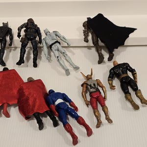 May include: A collection of superhero action figures in various poses. The figures are in different colors, including black, blue, red, gold, and gray. Some figures have capes. The figures are on a white surface.